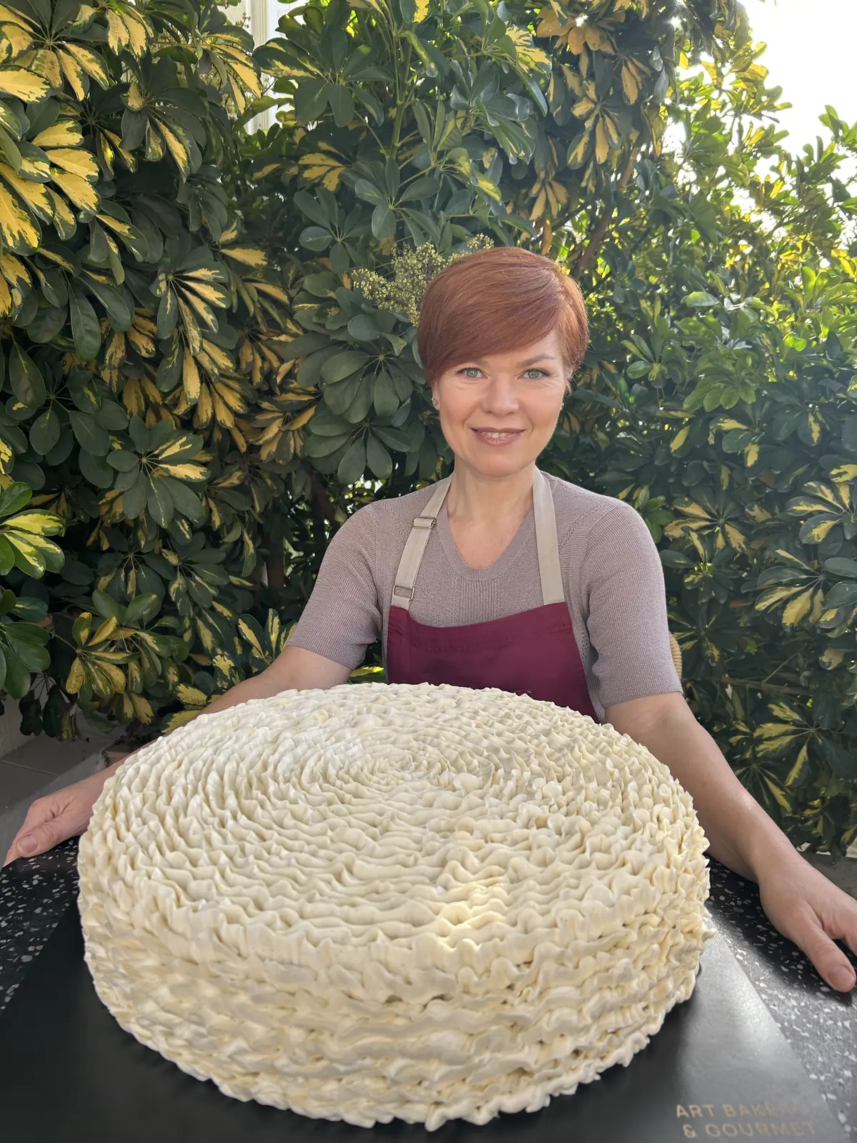 Tanya Novak smiling in her apron behind a large cream-covered celebration cake